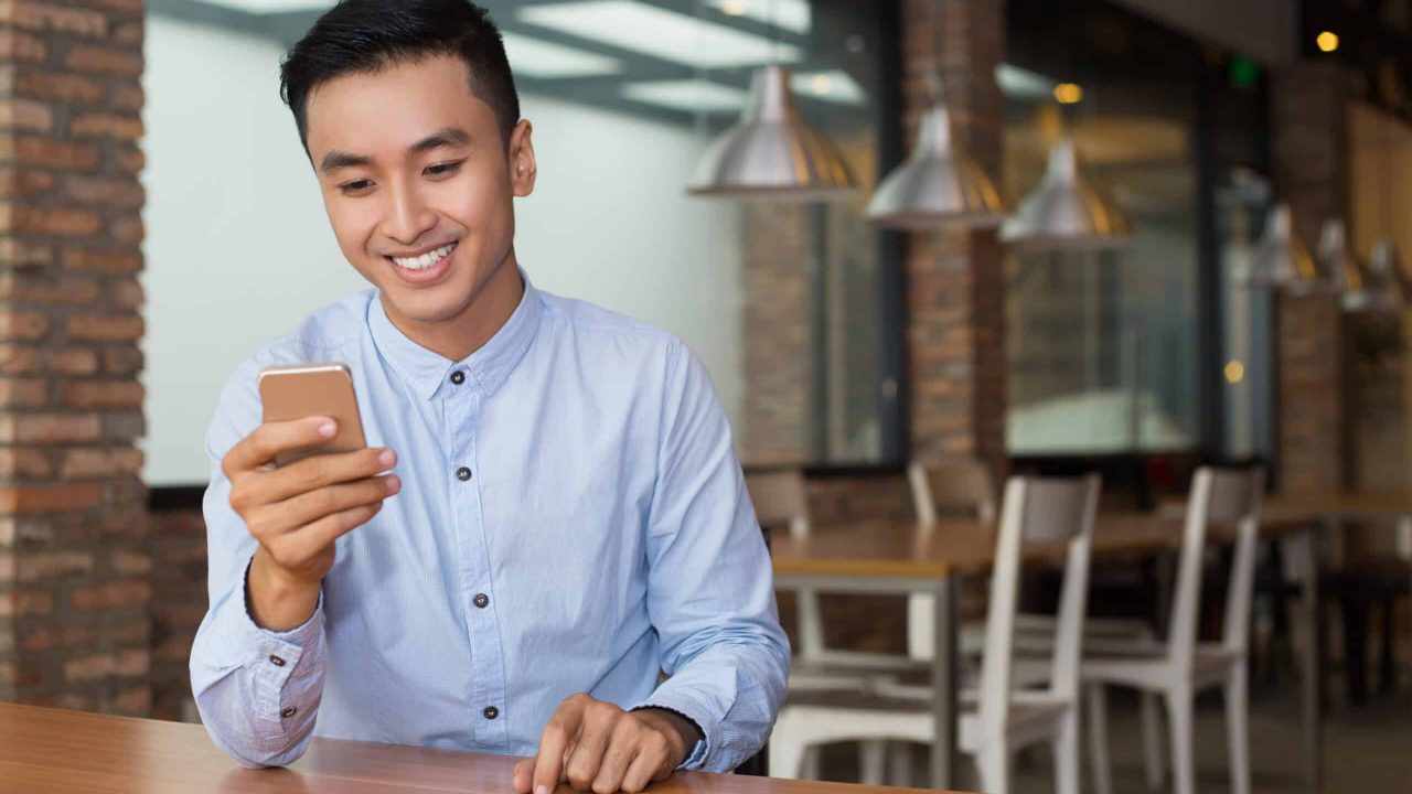 Closeup portrait of smiling young Asian man using smartphone and sitting at empty table with blurred cafe interior in background